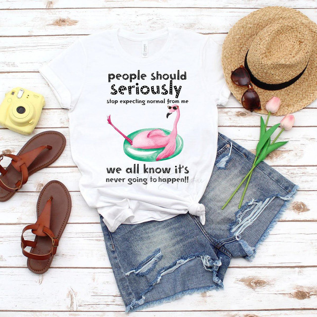 People Should Stop Expecting Normal From Me Flamingo Sublimation Transfer, humorous printed T-shirt with a pink flamingo wearing sunglasses, laid out on denim shorts with summer accessories like straw hat, sunglasses, tulips, sandals, and a yellow instant camera, on a wooden surface.