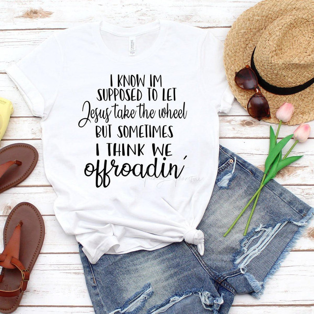 Jesus Take The Wheel Sublimation Transfer, white T-shirt with black and cursive text, laid flat on a white wooden surface with accessories including a straw hat, sunglasses, pink tulips, and casual sandals.