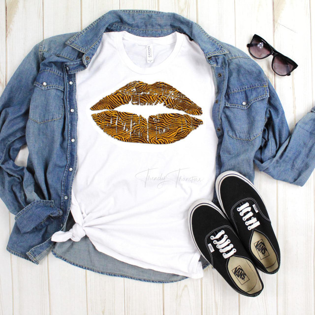 Tiger Animal Print Lips Sublimation Transfer, a graphic print of tiger-striped lips on a white T-shirt paired with a denim shirt, black Vans sneakers, and sunglasses laid on a wooden surface.