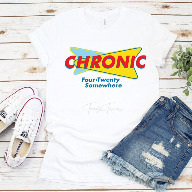 Chronic Sublimation Transfer, graphic design on a white T-shirt with the text 'Chronic' in bold red and yellow colors and 'Four Twenty Somewhere' below, laid out flat on a wooden surface with white sneakers and denim shorts.