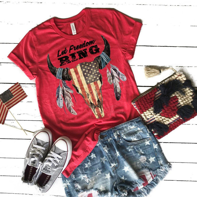 Patriotic Skull Screen Print Heat Transfer, red t-shirt with a skull design featuring an American flag pattern and the words "Let Freedom Ring". The outfit is styled with distressed denim shorts with star patterns, white sneakers with black stripes, and patriotic accessories including a flag pin and a woven clutch with fringe accents.