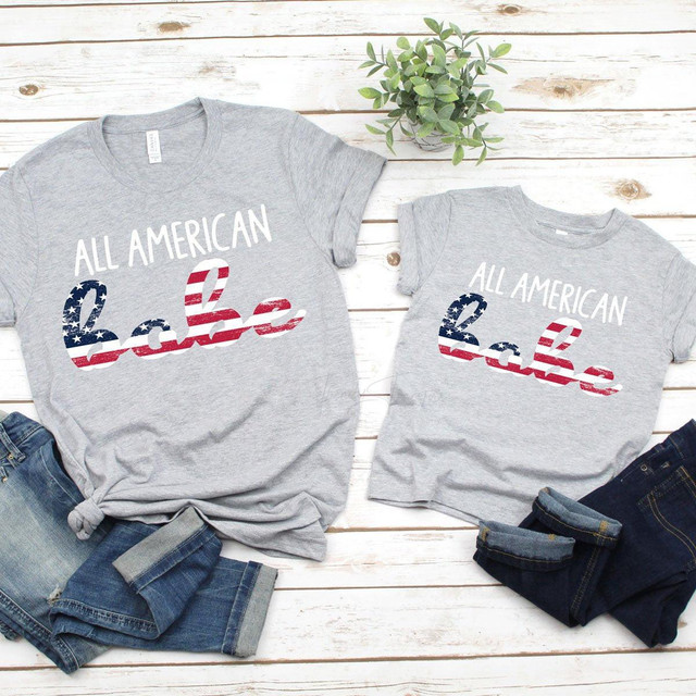 All American Babe ADULT Screen Print Heat Transfer, grey t-shirts with patriotic American flag design and text 'All American Babe' on a wooden background with folded jeans and a small green plant.