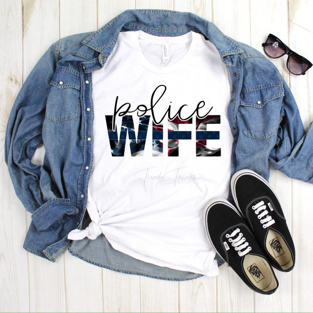 Police Wife Blue Line Flag Sublimation Transfer, a t-shirt with a design featuring an American flag motif and the words "police wife" printed on it, laid flat on a wooden surface with a denim shirt, black sunglasses, and black Vans sneakers around it.