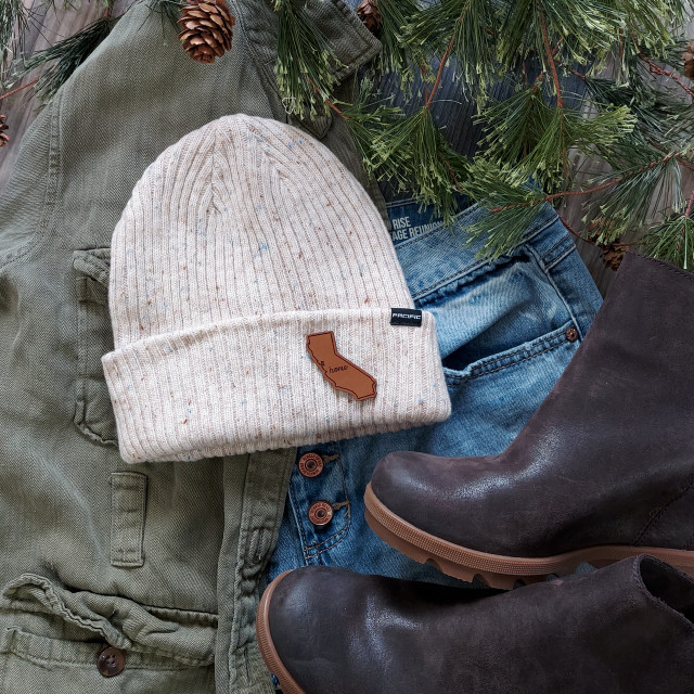 New Jersey CUSTOM Patch, beige knitted hat with a brown outline patch of the state of New Jersey with the word 'home' on it, laid on casual clothing and surrounded by pine branches and pinecones.