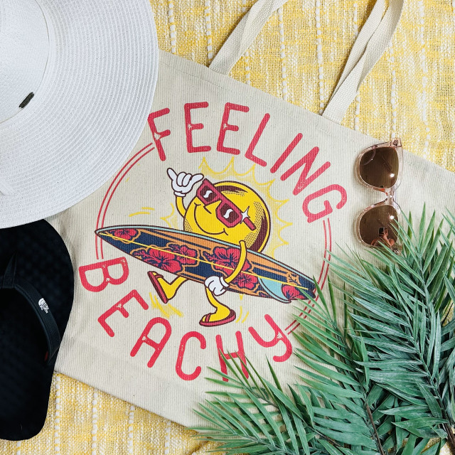 Feeling Beachy Surfboard Screen Print Heat Transfer, colorful beach-themed printed fabric with a surfing sun character wearing sunglasses, holding a surfboard, on a tote bag, surrounded by accessories like sunglasses and plants.
