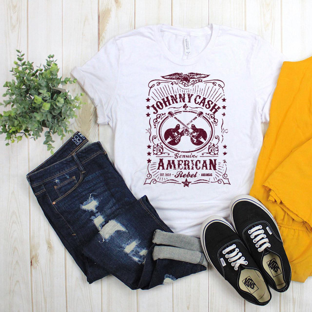 Johnny Cash Guitar Fan Art Label Red Glitter Effect Sublimation Transfer T-shirt, displayed on a flat lay with jeans, sneakers, yellow garment, and a green plant on a wooden background.