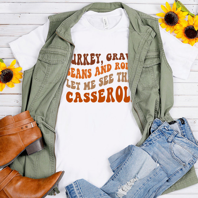 Turkey Gravy Casserole Sublimation Transfer, white t-shirt with brown and orange text design, laid flat with denim jeans, green vest, brown boots, and sunflower decorations on white wooden background.