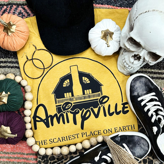 Amityville Screen Print Heat Transfer, yellow fabric with the Amityville haunted house logo and text, surrounded by pumpkins, skull, sneakers, earrings, and wooden beads.