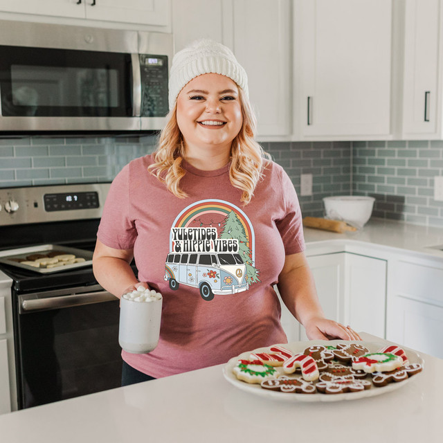 Yuletides & Hippie Vibes DTF Transfer, colorful retro-style design featuring a van, a rainbow, trees, and festive elements on a woman wearing a matching T-shirt in a modern kitchen setting.