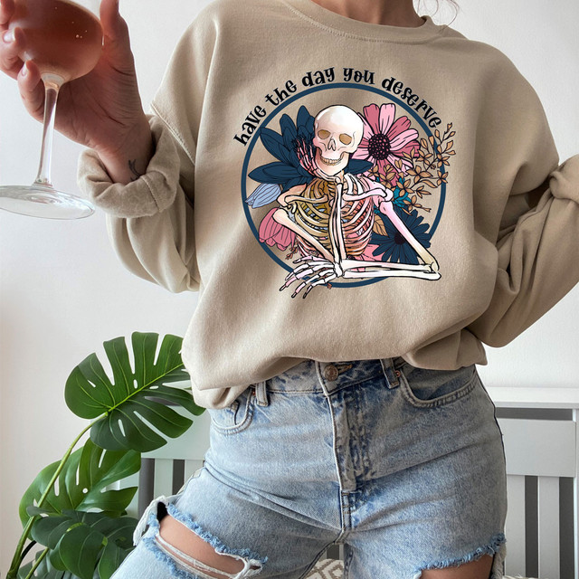 Have The Day You Deserve DTF Transfer, a colorful graphic design featuring a skeleton with flowers and the inspirational phrase on a beige sweatshirt.