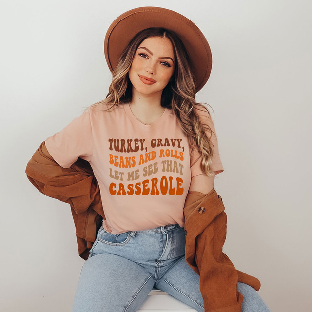 Let Me See That Casserole DTF T-shirt, woman wearing a stylish graphic T-shirt with playful text about turkey gravy, beans, rolls, and casserole, paired with a brown hat and a brown jacket.
