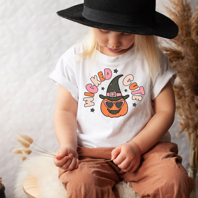 Wicked Cute Witch YOUTH Screen Print Heat Transfer, a young girl wearing a white T-shirt with a Halloween-themed pumpkin and witch hat design, and a black witch hat.