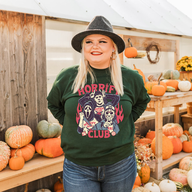 Horrify Club DTF Transfer, Halloween-themed graphic design of horror characters including a person with a ghost mask and a person with a scary mask, on a dark green sweatshirt worn by a woman at a pumpkin patch.