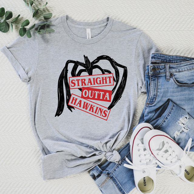 Product Straight Outta Hawkins DTF Transfer, graphic T-shirt with an illustration of an apple and the text "Straight Outta Hawkins" in red and black, laid flat with denim jeans and white sneakers, on a textured white background with a green leafy branch.