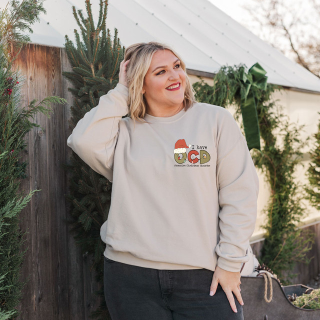 I Have OCD POCKET Screen Print Heat Transfer, a woman wearing a beige sweatshirt with holiday-themed OCD text and Christmas hat design, outdoors in a festive setting.