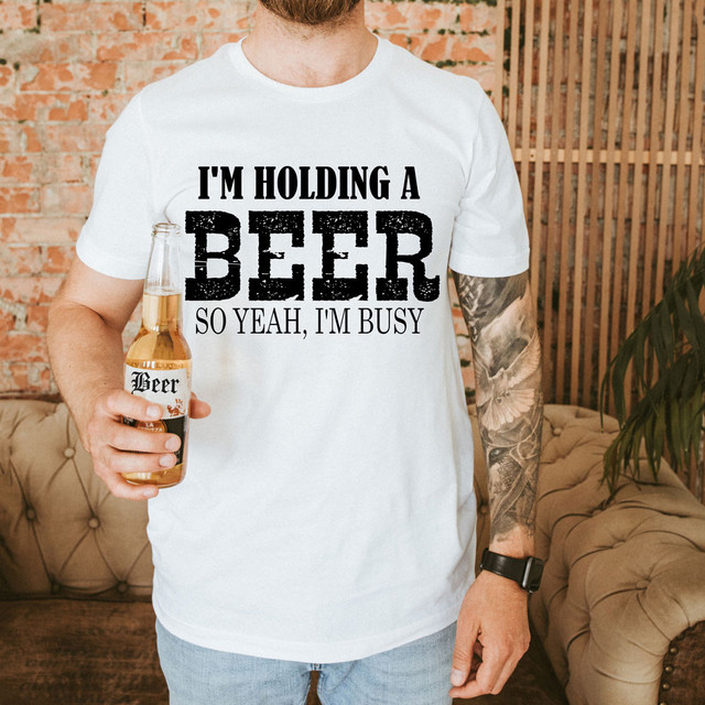 Product - I'm Holding A Beer So Yeah, I'm Busy Screen Print Heat Transfer - Man wearing a white T-shirt with humorous beer-themed print, holding a beer bottle, in a cozy indoor setting.