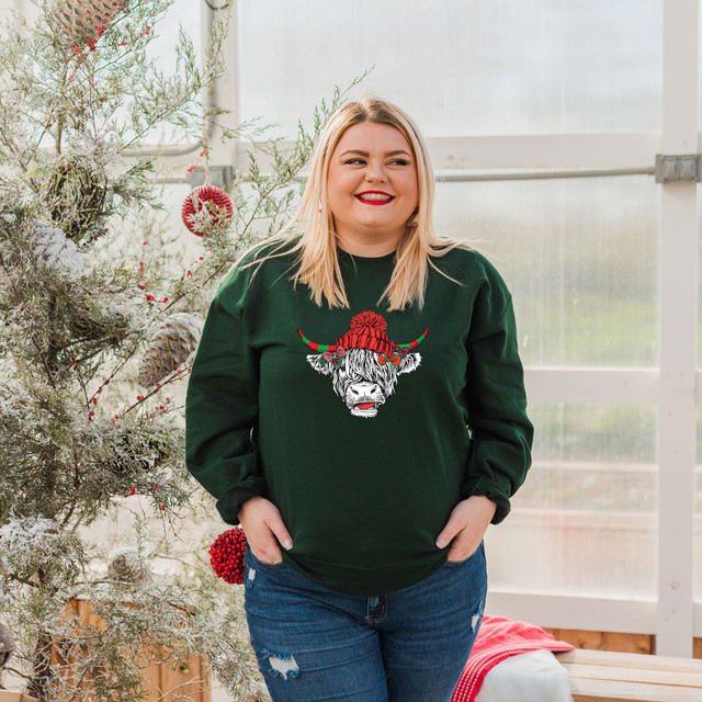 Christmas Shaggy Cow Hat DTF Transfer, a close-up of a cheerful woman wearing a green Christmas sweatshirt with a shaggy cow face design topped with a red Christmas hat and festive horns, in a holiday decorated setting.