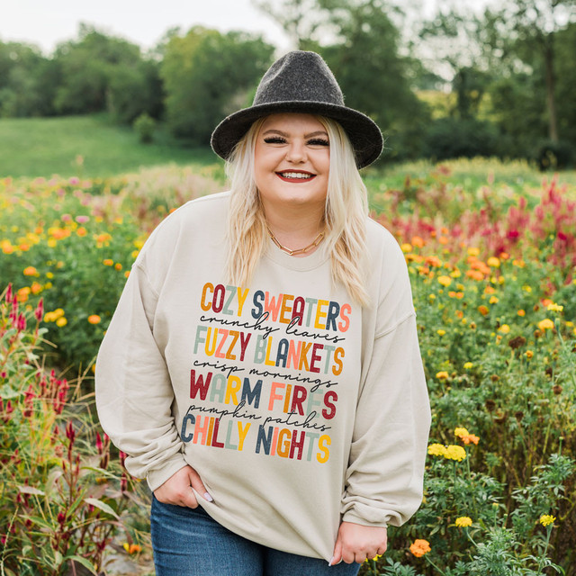 Cozy Sweaters Crunchy Leaves DTF Transfer, woman wearing a fall-themed sweatshirt with colorful text, standing in a vibrant flower field with trees in the background.