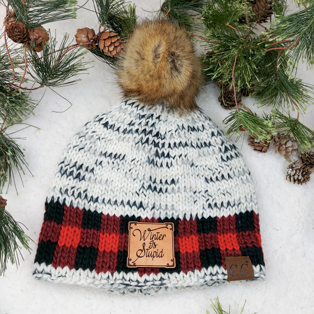Winter Is Stupid TAN Leather Patch, cozy knit beanie with faux fur pom-pom, surrounded by pine branches and pinecones in a winter setting.