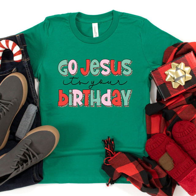 Go Jesus It's Your Birthday DTF Transfer, colorful t-shirt with holiday-themed text and designs, surrounded by wrapped gift box, plaid scarf, red knit gloves, shoes, and Christmas decorations.
