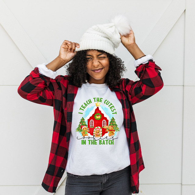 I Teach The Cutest Cookies In The Batch DTF Transfer, woman wearing a holiday-themed shirt with a gingerbread house and Christmas decorations with playful phrase, standing against a white background