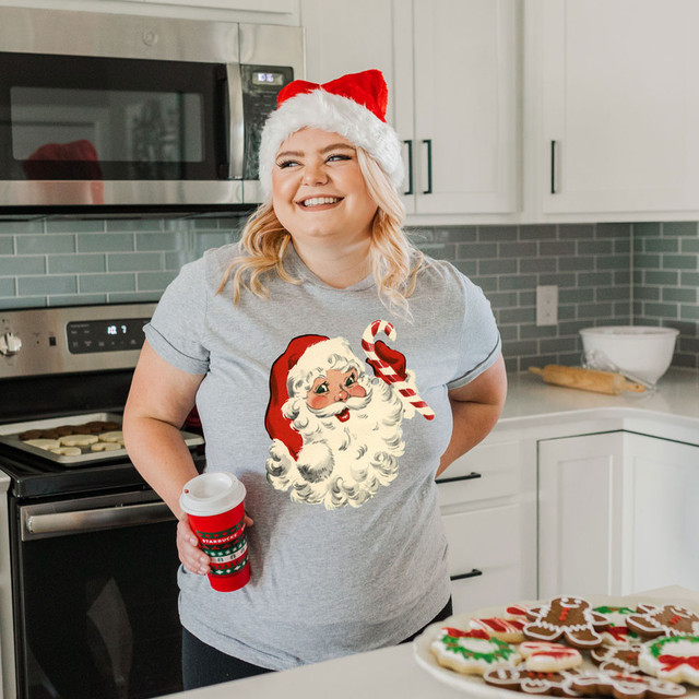 Vintage Santa Red Hat DTF Transfer, woman wearing a Santa hat and a T-shirt with a vintage Santa design holding a holiday coffee cup in a modern kitchen.