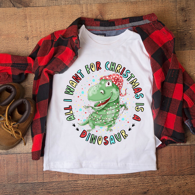 All I Want For Christmas Is A Dinosaur DTF Transfer T-shirt, featuring a cute cartoon dinosaur wearing a Christmas hat with festive decorations around it.