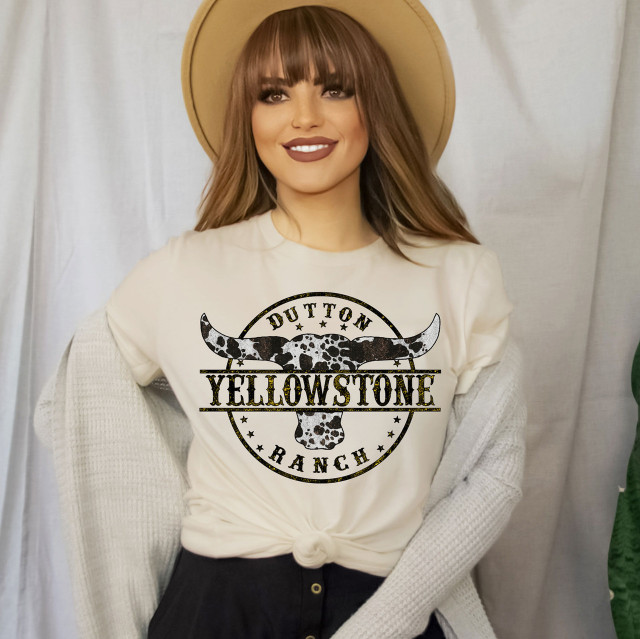 Dutton Ranch Screen Print Heat Transfer, woman wearing a white t-shirt featuring a black and white cow skull with the text Dutton Yellowstone Ranch, with a beige hat and cozy cardigan.