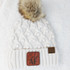 Cold AF Leather Patch, close-up image of a stylish winter hat with a faux fur pom-pom and knit pattern, featuring a leather patch with the brand's logo.
