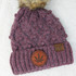 Product A Toke A Day Keeps The Bullshit Away Leather Patch, a knitted winter hat with a brown leather patch featuring the text and a cannabis leaf logo, and a small black leather logo tag with the initials C.C.