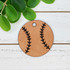 Personalized Leather Baseball Softball Keychain, round leather keychain with black baseball stitch design and a small hole for attachment on a white wooden surface with green leaves nearby.