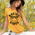 Racing Vibes Happy Face DTF Heat Transfer, a smiling face with checkered sunglasses and the words 'Racing Vibes' on a yellow t-shirt worn by a woman outdoors amidst yellow flowering bushes.