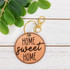 Home Sweet Home Leather Keychain, round leather keychain with 'Home Sweet Home' text in black, attached to a gold keyring and clasp, on a white wooden surface with green leaves.