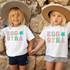 Eggstra Chenille 'look' DTF Heat Transfer, two young girls wearing white T-shirts with colorful embroidered 'Eggstra' designs while holding hands and standing against a stone wall.