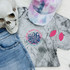 Afterlife Support Group POCKET Screen Print Heat Transfer, colorful apparel and accessory items including a gray T-shirt with the design, pink and black striped earrings, a pastel tie-dye cap, a skull prop, and denim jeans.