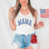 Mama Stars DTF Heat Transfer, close-up of a woman wearing a white tank top with 'MAMA' in star-spangled text, holding an American flag, with patriotic accessories and distressed denim shorts.