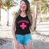 Lifeguard Pink DTF Heat Transfer, a woman wearing a black tank top with the word 'LIFEGUARD' and a pink cross graphic, on a beach with palm trees.