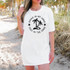 Drink In My Hand Toes In The Sand DTF Heat Transfer, a woman wearing a white t-shirt with a beach-themed graphic design featuring palm trees and the phrase 'Drink In My Hand, Toes In The Sand' on a sandy beach with palm trees in the background.