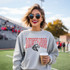 Mustangs *CUSTOM COLOR OPTIONS* DTF Heat Transfer, a woman wearing a grey sweatshirt with a Mustangs logo and holding a coffee, at an outdoor sports event.