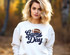 Navy Game Day Football Chenille Patch, front view of a young woman wearing a white sweatshirt with a colorful embroidered football and 'Game Day' text on it, outdoors in autumn.