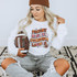 Friday Nights Stadium Lights DTF Heat Transfer, woman wearing sweatshirt with football-themed design, sitting on rug with football in hand.
