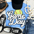 Softball White Game Day Chenille Patch, embroidered patch with a softball and the words 'Game Day' on a blue T-shirt surrounded by sneakers, a black cap, a beaded necklace, and denim jeans.