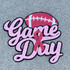 Breast Cancer Game Day Chenille Patch, pink and red embroidered patch with a football, pink ribbon, and the words "Game Day" on a gray surface.