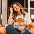 Fall In Love With Learning DTF Heat Transfer, a woman wearing a beige T-shirt with a colorful retro design and daisies, sitting among pumpkins, smiling outdoors during fall.