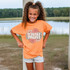 Mini Boho Floral DTF Heat Transfer, a girl wearing an orange t-shirt with a floral design and the word 'MINI' printed on it, outdoors by a lake with trees in the background.