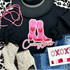 Cowboy Lover Sequin Patch, pink and red cowboy boots with heart accents and the word Cowboy Lover embroidered below, on a black sweatshirt surrounded by pink bead necklace, leopard print boots, denim jeans, and Valentine's Day themed decorations.