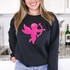 Pink Cupid Sequin Patch, a pink shiny sequin patch of Cupid with a bow and arrow featuring wings and curly hair, on a black sweatshirt.