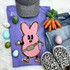 Trendy Pink Bunny Chenille Patch, pink bunny character with big eyes and a bag, on a purple shirt with other fashion accessories and decorative items around.
