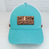 Shit Show Supervisor Leather Hat Patch, turquoise trucker cap with pink and brown detailing, and a beige patch with black text that reads 'Shit Show Supervisor'.