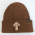 Big Mushroom Leather Hat Patch, brown knitted beanie hat with speckled yellow accents and a mushroom patch decoration.